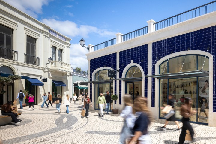 Outdoor shopping area with people walking by stores, including a Furla shop with blue tiled facade.