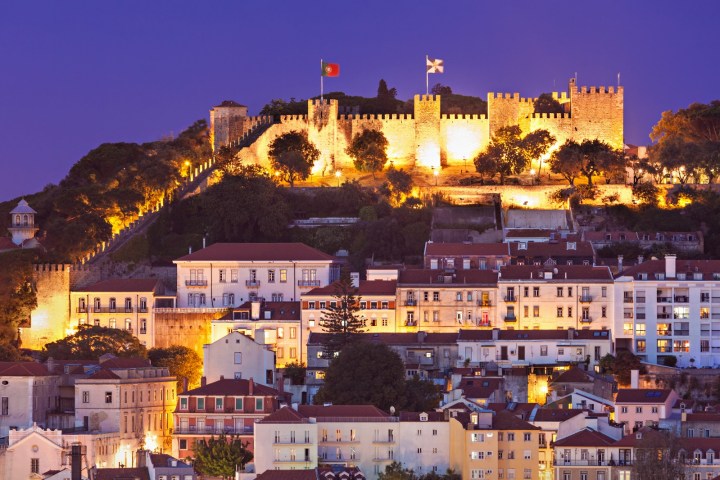 Illuminated castle on a hill with city buildings in foreground at dusk.