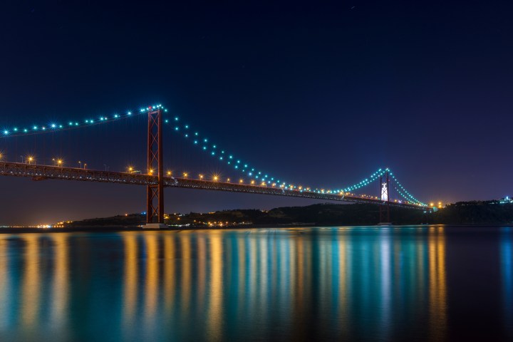 Illuminated suspension bridge over calm water at night with city lights in the background.