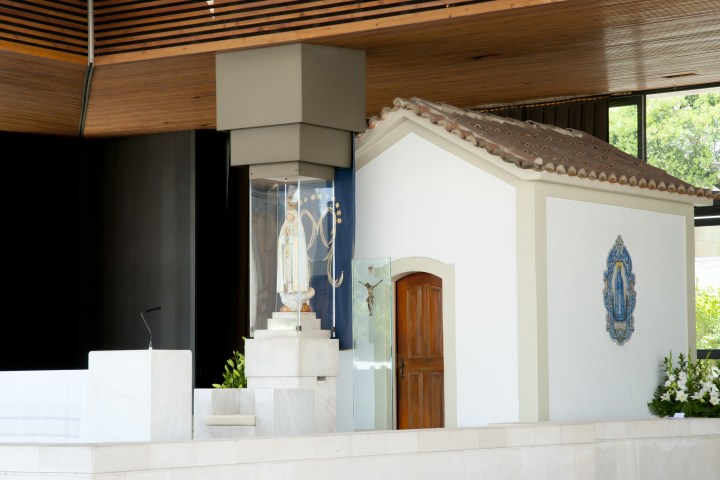 Chapel with a statue of Virgin Mary in a glass case, wooden door, and religious emblems on white walls.