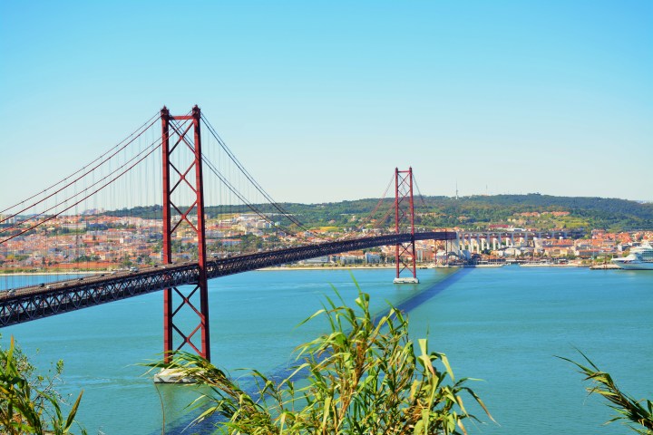 Red suspension bridge over a blue river with a city in the background on a clear day.