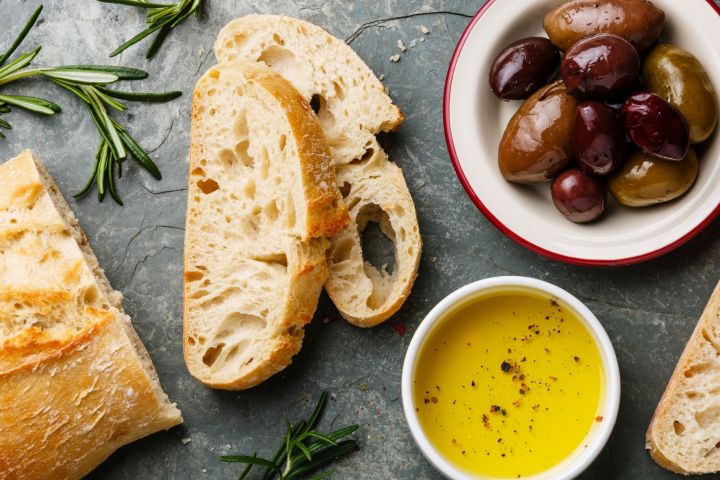Slices of bread, olives in a bowl, rosemary, and olive oil with pepper on a dark surface.