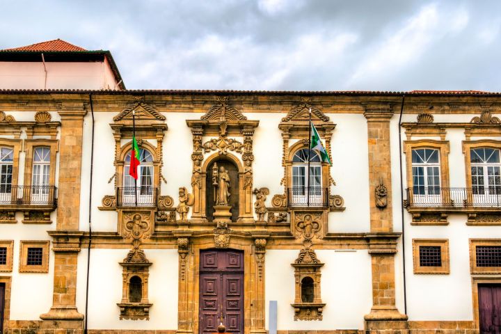 Ornate historic building facade with flags and a fountain in foreground under a cloudy sky.