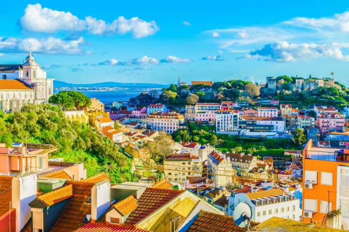 Scenic view of Lisbon with colorful buildings, red roofs, greenery, and distant water under a blue sky with clouds.