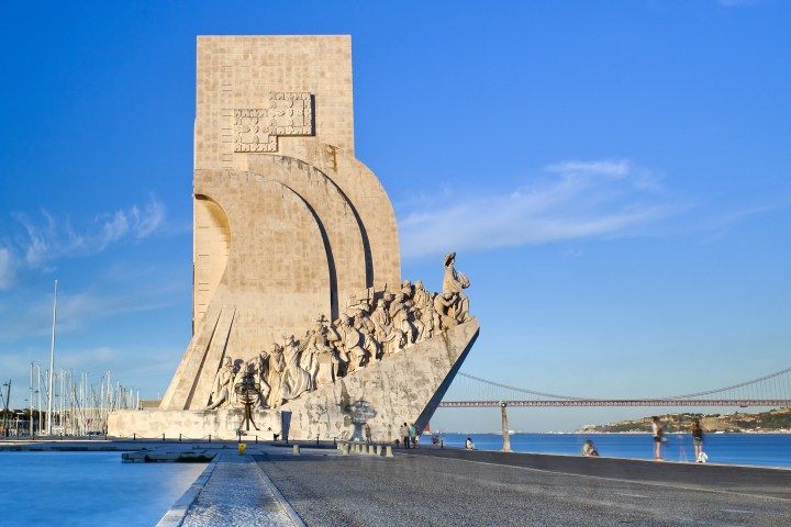 Monument with sculptures of figures by a waterfront, clear sky, and bridge in the background.