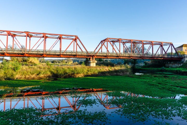 Red steel bridge over a river with green plants, reflecting in the water.