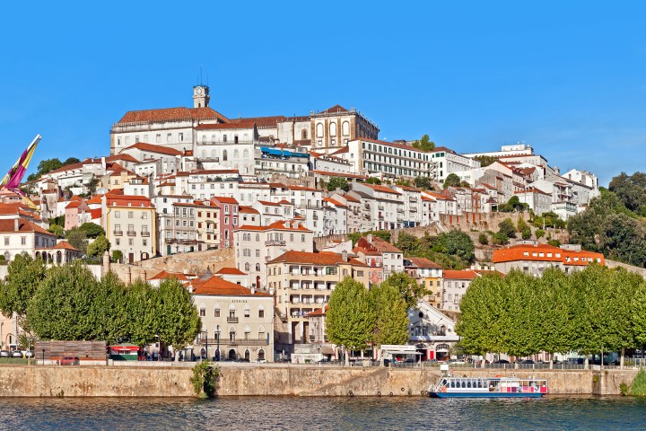 Scenic view of Coimbra, Portugal with hillside buildings and a river in the foreground under a clear blue sky.