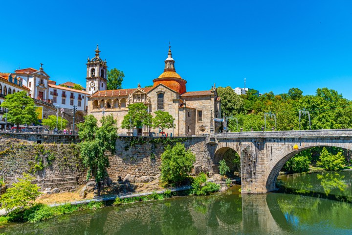 Historic stone bridge and church with red roof over a river, surrounded by trees and blue sky.