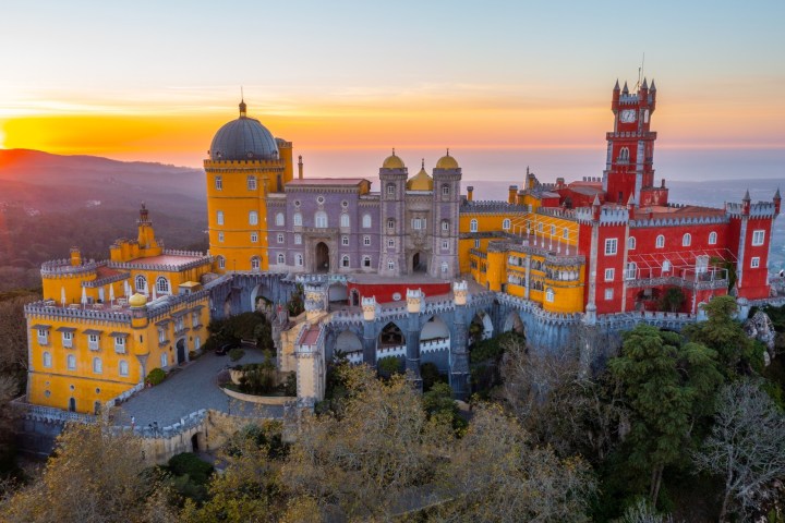 Colorful palace with domes and towers at sunset, surrounded by trees and hills.