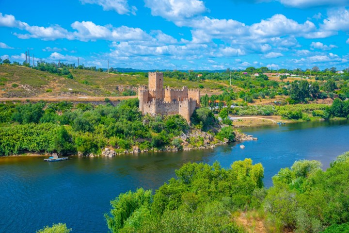 A medieval castle on a river island, surrounded by lush green landscape under a blue sky with clouds.