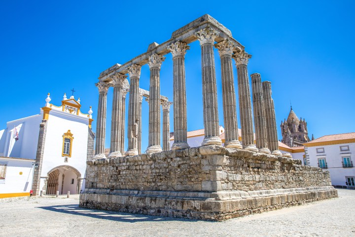 Ancient Roman temple ruins with stone columns near a white church under a clear blue sky.