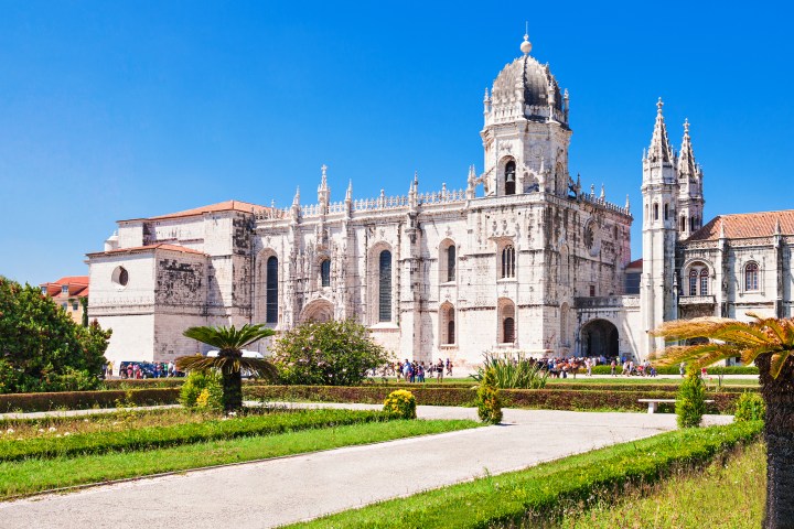 Historic stone building with ornate spires and garden in foreground on a clear day.