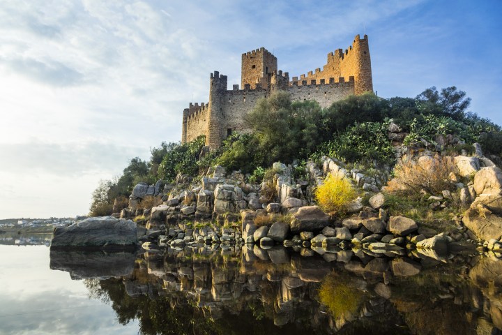 Stone castle on a rocky hill reflected in calm water under a blue sky.