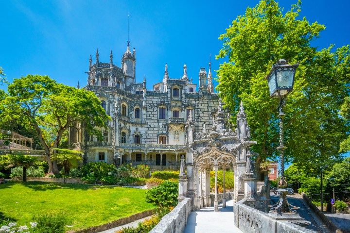 Gothic-style castle with ornate facade surrounded by trees and garden under a sunny blue sky.