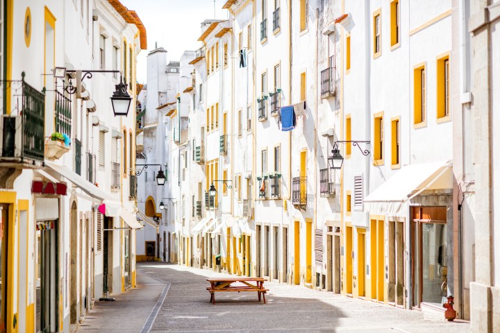 Narrow European street with white buildings, yellow accents, and hanging laundry, featuring vintage lamps.