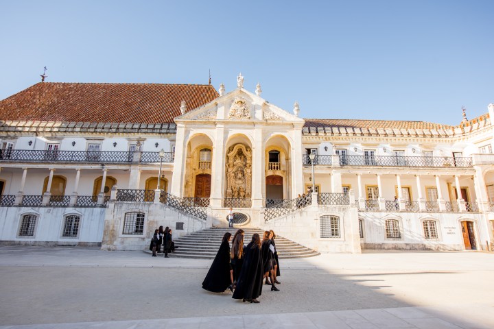 Group of people in black cloaks walking in front of a historical building with a large courtyard.