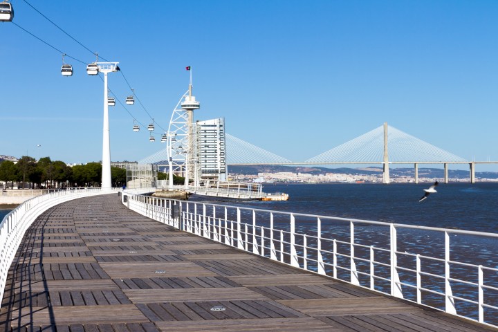 Wooden walkway by river with cable cars, bridge, and tower in background.
