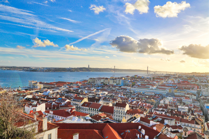 Scenic view of a city with red rooftops, river, and a distant suspension bridge under a partly cloudy sky.