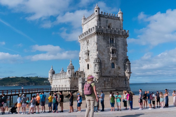 People line up near a historic stone tower by the sea on a sunny day.