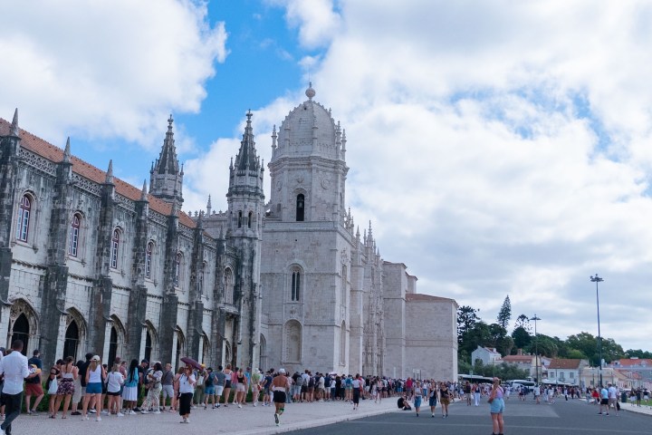 Historic building with ornate towers and a long line of people outside under cloudy skies.