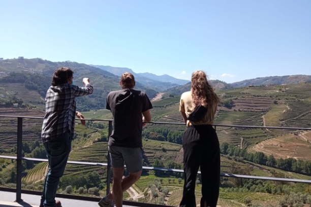 Three people on a balcony overlook terraced hills under a clear blue sky.
