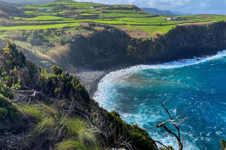Cliffside view of rocky shore with waves, green fields, and distant hills under a blue sky.