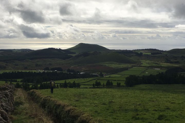 Person walking on grassy hill, overlooking distant hills and cloudy sky.