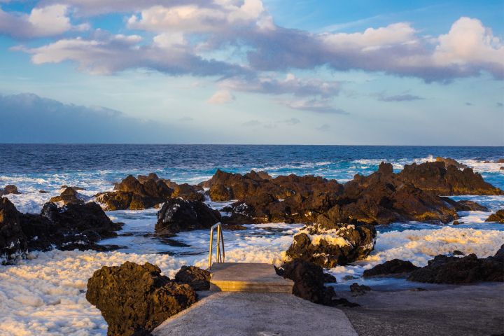 Rocky coastline with ocean waves and clouds under a blue sky.