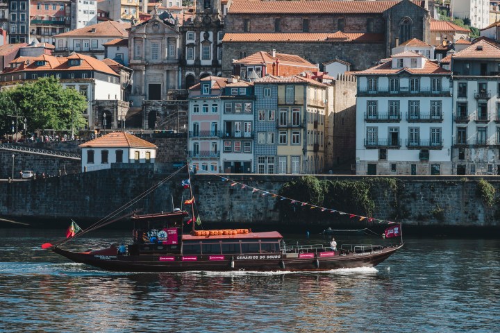 A traditional boat sails on a river with colorful buildings and a church in the background.