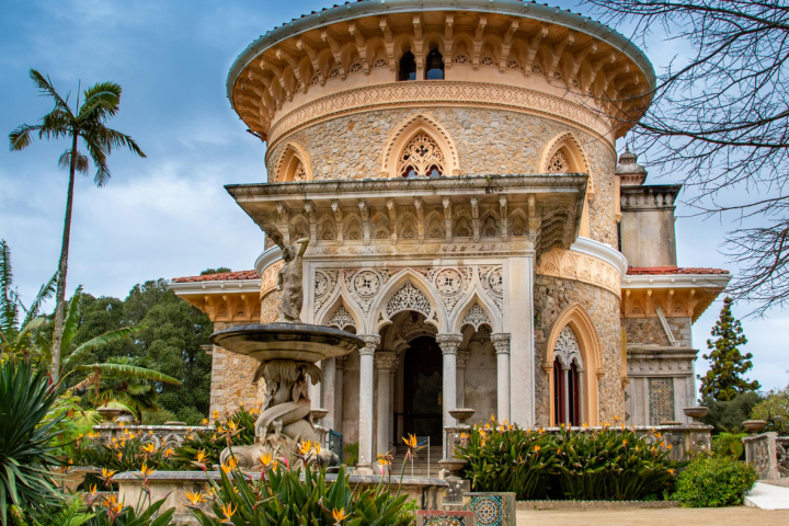 Ornate round building with arches, surrounded by gardens and palm tree under a cloudy sky.
