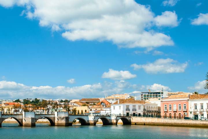 Stone bridge over calm water with colorful buildings and blue sky.