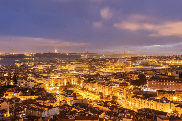 Cityscape of Lisbon at dusk with illuminated buildings and a distant bridge under a cloudy sky.