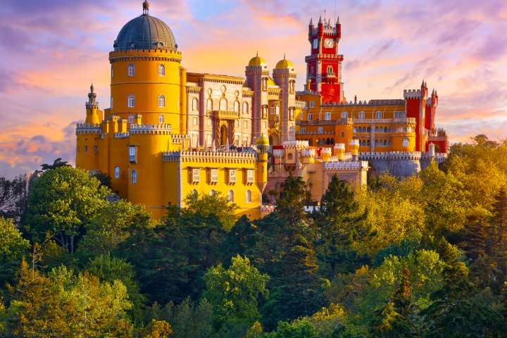 Colorful castle with domes and towers surrounded by trees against a sunset sky.