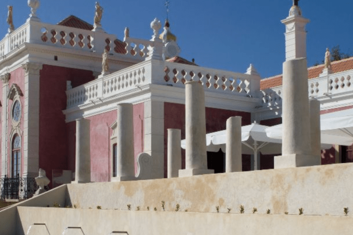 Historical building with pink facade, ornate pillars, and fountains under a clear blue sky.