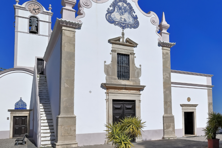 White church with clock tower, decorated tiles, and a cross, set against a clear blue sky.