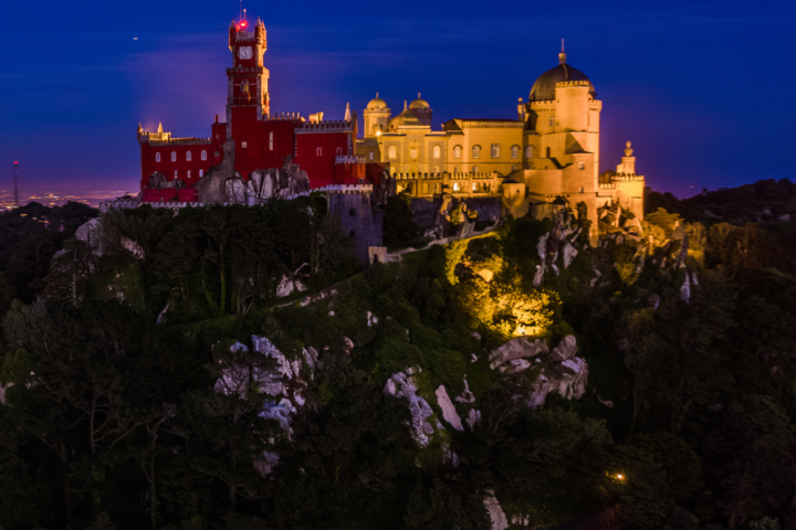 Illuminated castle on hill with red and yellow buildings at dusk.