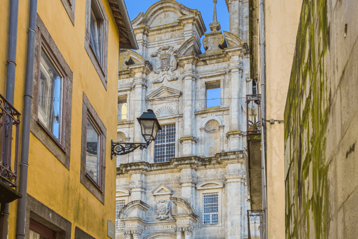 Narrow street view of ornate historic church facade with cross on top, framed by yellow walls and blue sky.