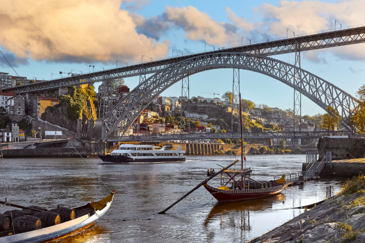 Bridge over river with boats; cityscape in background under a cloudy sky.