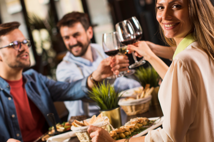 Three people toasting with wine glasses around a table with food and plants in a cozy restaurant setting.