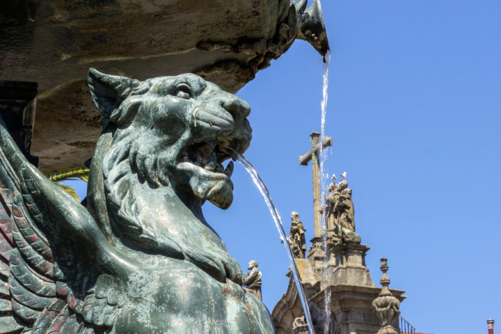 Stone griffin fountain with water spouting, church facade in background under clear blue sky.