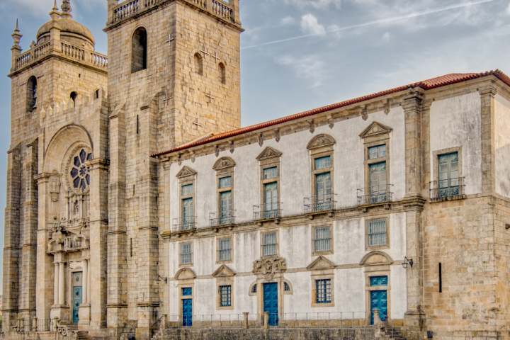 Historic stone cathedral with two towers and adjacent white building, under a partly cloudy sky.
