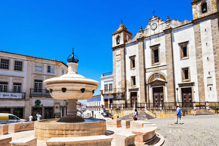 Open plaza with historic church and stone fountain under a clear blue sky.