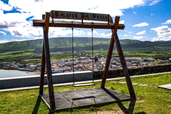 Wooden swing overlooking a coastal town and green hills under a blue sky with clouds.