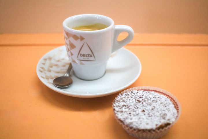 Espresso in a white cup on saucer with spoon, next to a powdered sugar-topped muffin on orange table.