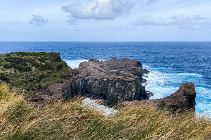 Rocky coastline with grassy cliffs overlooking a blue ocean under a partly cloudy sky.