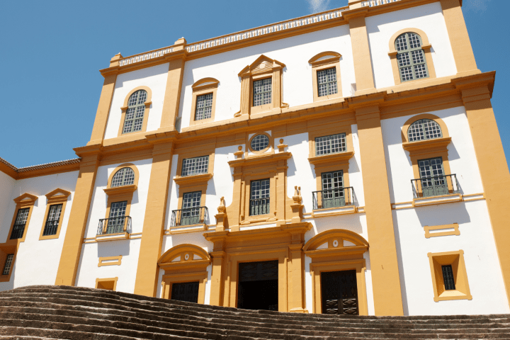 White and yellow baroque-style building with arched windows and stone steps under blue sky.