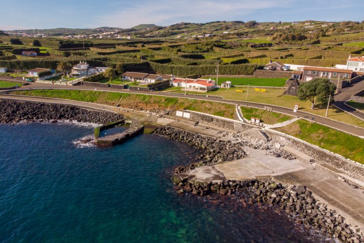 Coastal view with concrete dock, rocky shore, green fields, and houses.