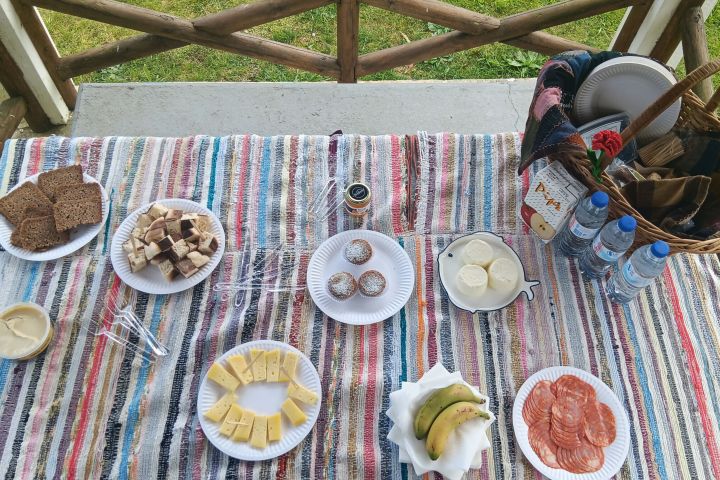 Table with assorted cheeses, bread, bananas, salami, muffins, and drinks on a striped cloth.
