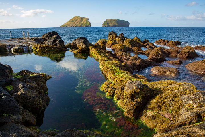 Rocky coastal landscape with tidal pool and distant islets under a clear blue sky.