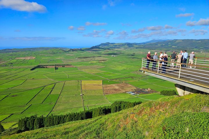Tourists on a viewing platform overlook a vast green patchwork of fields under a blue sky.